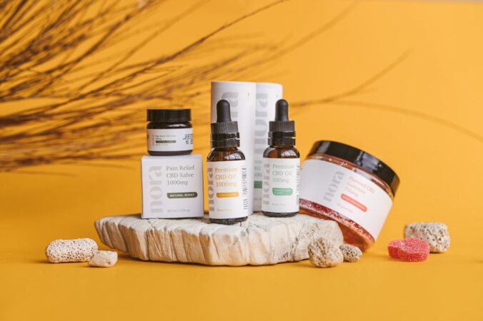 Nora brand CBD products including oil drops, pain relief salve, and gummies displayed on stone against yellow background